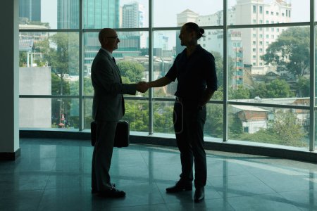 Téléchargez les photos : Aperçu de deux partenaires commerciaux qui se serrent la main contre une grande fenêtre du centre de bureau tout en se félicitant pour un nouvel accord - en image libre de droit