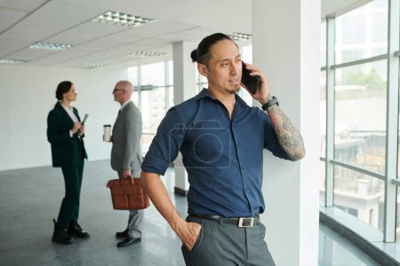 Foto de Joven asiático solopreneur en camisa y pantalones hablando por teléfono móvil mientras está de pie delante de la cámara en el centro de oficina - Imagen libre de derechos