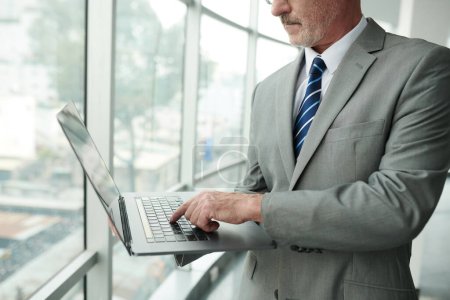 Photo for Cropped shot of mature male solopreneur in elegant grey suit pressing key of laptop keyboard while standing by large window in office center - Royalty Free Image