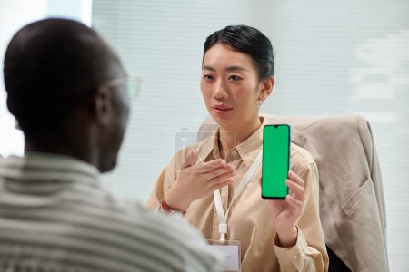 Téléchargez les photos : Gestionnaire de banque souriant montrant smartphone client avec écran vert, expliquant comment utiliser l'application bancaire - en image libre de droit