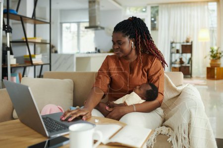 Foto de Mujer negra sentada en el sofá con el bebé recién nacido en el ordenador portátil al tener una reunión en línea - Imagen libre de derechos