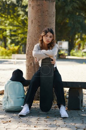 Téléchargez les photos : Portrait d'adolescente sérieuse avec planche à roulettes assise sur un banc sous un arbre - en image libre de droit