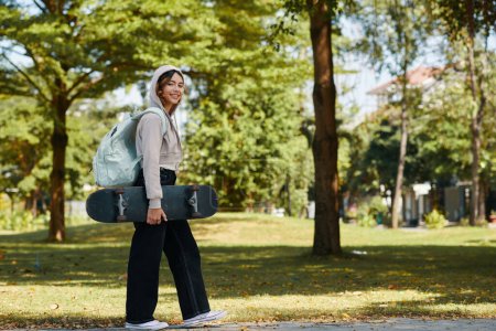 Téléchargez les photos : Portrait de fille gaie avec planche à roulettes marchant à la maison à travers le parc après les cours - en image libre de droit