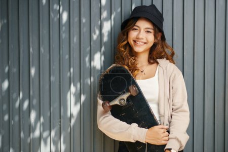 Téléchargez les photos : Joyeux adolescent fille dans le seau chapeau debout au mur de garage avec planche à roulettes dans les mains - en image libre de droit