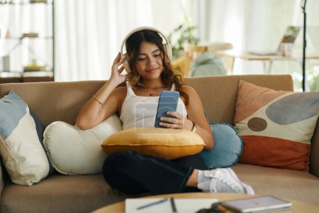 Photo for Smiling girl sitting on couch and watching music videos on smartphone - Royalty Free Image
