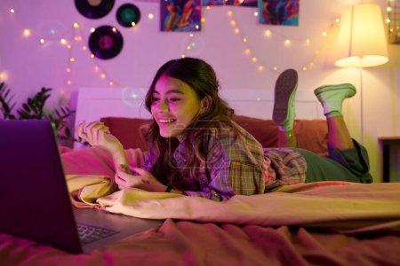 Téléchargez les photos : Adolescente couchée sur le lit en face de l'ordinateur portable ouvert lorsque l'appel vidéo ami - en image libre de droit