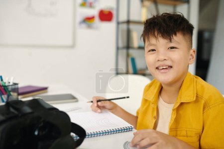 Foto de Retrato de colegial alegre sentado en el escritorio en clase y escribiendo en libro de texto - Imagen libre de derechos