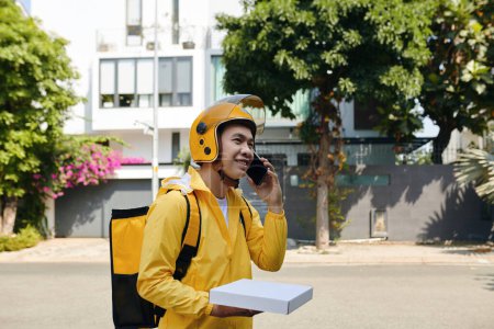 Foto de Repartidor en uniforme amarillo llamando al cliente para discutir dónde dejar la caja de pizza - Imagen libre de derechos