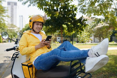 Téléchargez les photos : Courrier souriant assis sur scooter, se reposant entre les promenades, textos amis - en image libre de droit