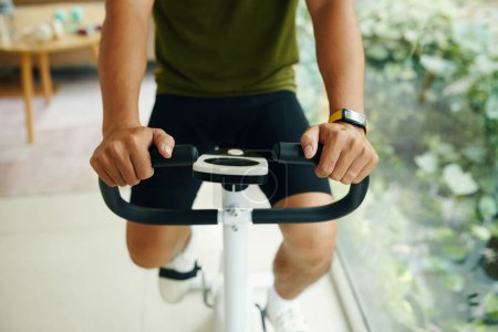 Téléchargez les photos : Image recadrée de l'homme portant un pisteur de forme physique lors de l'entraînement sur vélo d'appartement - en image libre de droit