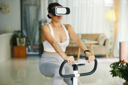 Téléchargez les photos : Fit femme portant casque de réalité virtuelle lors de l'équitation vélo d'appartement - en image libre de droit