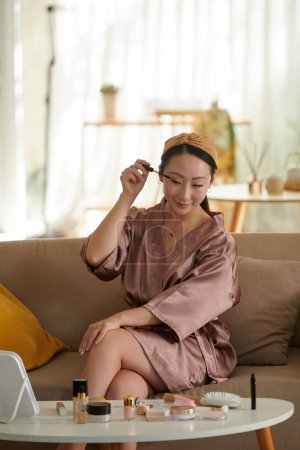 Smiling woman in satin robe applying mascara when getting ready in the morning