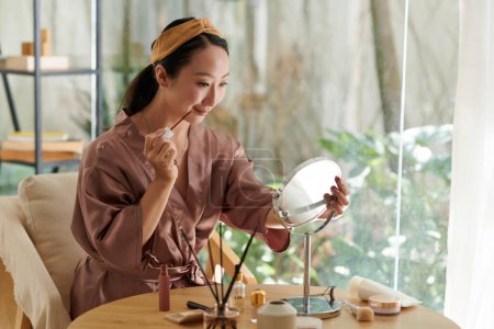 Téléchargez les photos : Sourire jeune femme chinoise appliquant le maquillage lors de la préparation pour la date ou l'événement - en image libre de droit