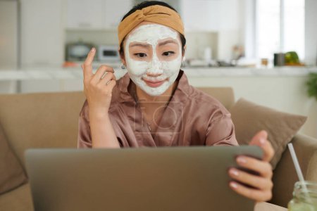 Téléchargez les photos : Femme souriante avec masque en argile purifiante à la lecture de l'article sur l'écran d'ordinateur portable - en image libre de droit