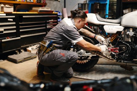Téléchargez les photos : Réparateur portant une ceinture-outil lors de la fixation de la moto dans le garage - en image libre de droit