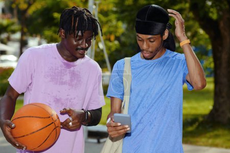 Verschwitzte Streetballspieler lesen Benachrichtigungen nach dem Spiel auf dem Smartphone