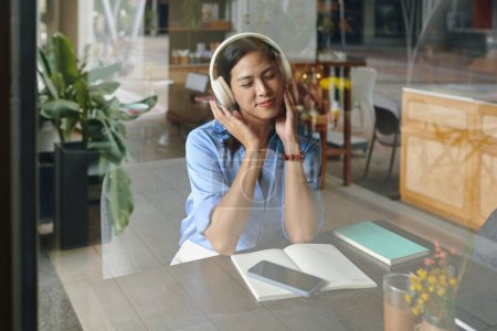 Téléchargez les photos : Femme heureuse assise à la table de café et profitant de la bonne musique dans les écouteurs - en image libre de droit
