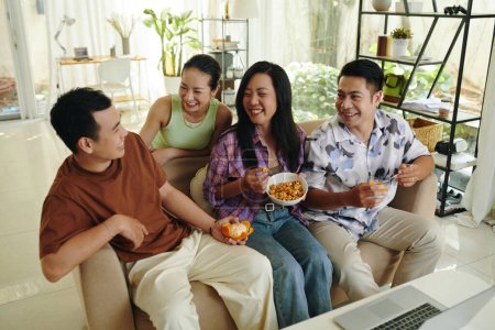 Téléchargez les photos : Groupe d'amis traînant à la maison, mangeant des collations et plaisantant autour - en image libre de droit