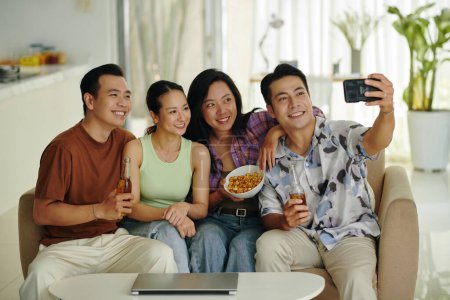 Téléchargez les photos : Groupe d'amis souriants assis sur le canapé à la maison et prenant selfie - en image libre de droit