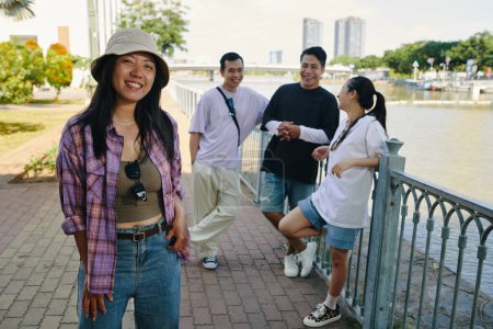 Téléchargez les photos : Souriant jeune femme vietnamienne traînant avec des amis sur le quai de la rivière - en image libre de droit