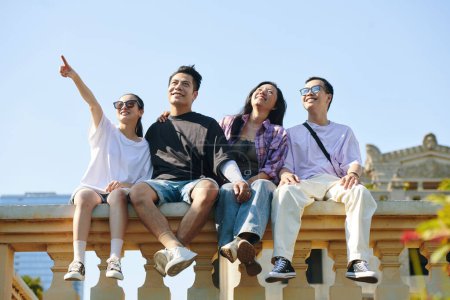 Téléchargez les photos : Jeunes gens joyeux assis sur la balustrade en pierre et profitant de la vue sur la ville - en image libre de droit
