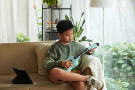 Téléchargez les photos : Pensive preteen garçon essayant de jouer ukulele à la maison - en image libre de droit