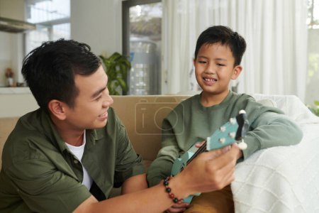 Téléchargez les photos : Souriant père enseignement fils jouer ukulele - en image libre de droit