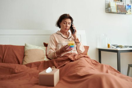 Photo for Sick woman sitting in bed in pajamas and calling doctor to ask what pills to tale - Royalty Free Image