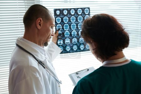 Foto de Neurocirujano examinando la radiografía cerebral del paciente cuando la enfermera escribe recomendaciones - Imagen libre de derechos