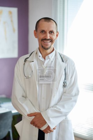 Portrait of happy experienced general practitioner in white coat standing in his office