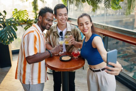 Téléchargez les photos : Souriant jeune femme prenant selfie avec des amis quand ils traînent dans le bar sur le toit - en image libre de droit