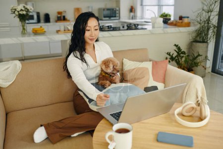 Téléchargez les photos : Chinois jeune femme assis sur le canapé avec petit chien et regarder un film sur ordinateur portable - en image libre de droit