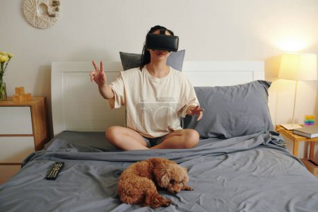 Photo for Young woman in homewear sitting on bed with her dog and playing game in vr glasses - Royalty Free Image