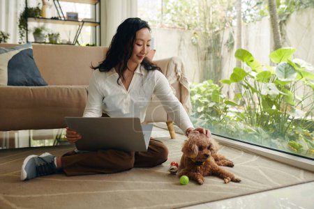 Photo for Young woman sitting on floor with opened laptop and petting dog - Royalty Free Image