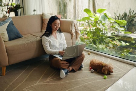 Photo for Young woman sitting on floor and working on laptop when her dog playing near by - Royalty Free Image