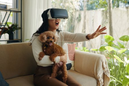 Téléchargez les photos : Jeune femme excitée tenant son chien en jouant au jeu vidéo dans des lunettes vr - en image libre de droit
