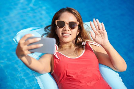 Téléchargez les photos : Femme souriante en maillot de bain rouge vidéo appelant des amis lors de la détente sur matelas gonflable - en image libre de droit