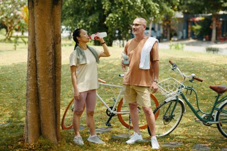 Active mature diverse couple standing under tree and drinking water after bicycle ride