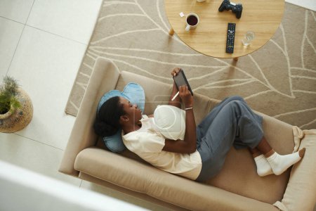 Foto de Tired Black girl lying on sofa and drawing sketches on tablet for design class - Imagen libre de derechos