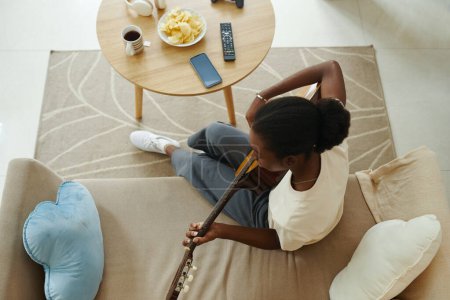 Photo for Black girl sitting on couch at home and playing guitar - Royalty Free Image