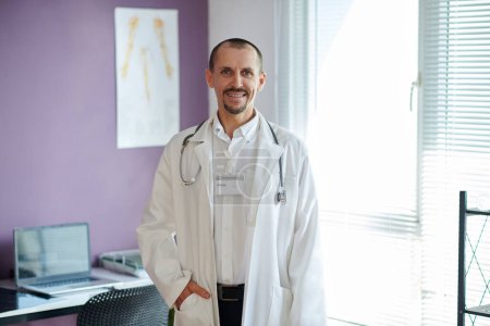 Retrato del doctor maduro sonriente usando bata de laboratorio de pie en su oficina