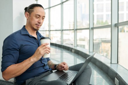 Téléchargez les photos : Homme d'affaires souriant boire du café et vérifier les e-mails sur ordinateur portable - en image libre de droit