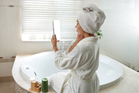 Téléchargez les photos : Femme en robe blanche assise sur le bord du bain et appliquant de la crème visage - en image libre de droit