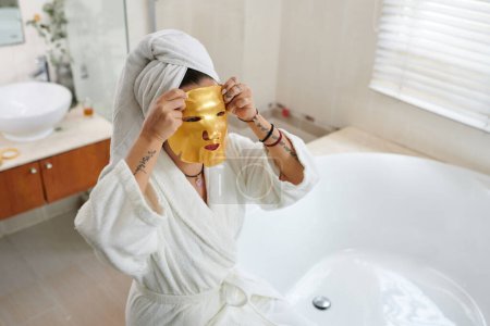 Téléchargez les photos : Femme appliquant un masque visage hydratant doré - en image libre de droit