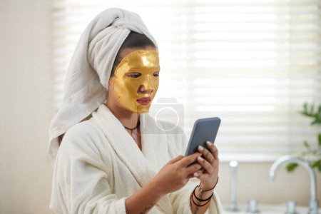 Photo for Portrait of woman with golden sheet mask on her face texting friends - Royalty Free Image