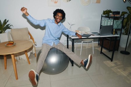 Photo for Happy Black entrepreneur bouncing on fitness ball when having break in office - Royalty Free Image