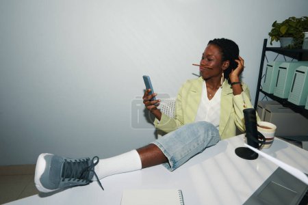 Geschäftsfrau sitzt mit Fuß auf Büroschreibtisch und macht Selfie mit Bleistift zwischen Lippen und Nase