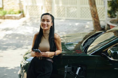 Portrait of happy Vietnamese young woman standing at her hybrid car, waiting for charging