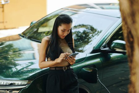 Young woman checking charging application on smartphone when standing at her electric car