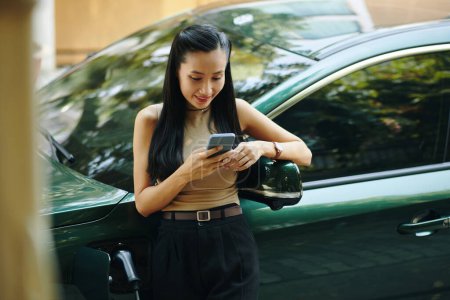 Elegant smiling young woman leaning on electric car, waiting for charging